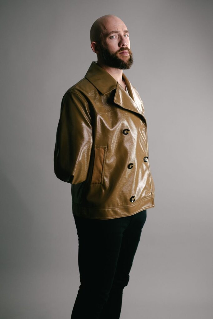 Dan a new model at Zebedee is an arm amputee, poses wearing a shiny brown jacker and black trousers. 